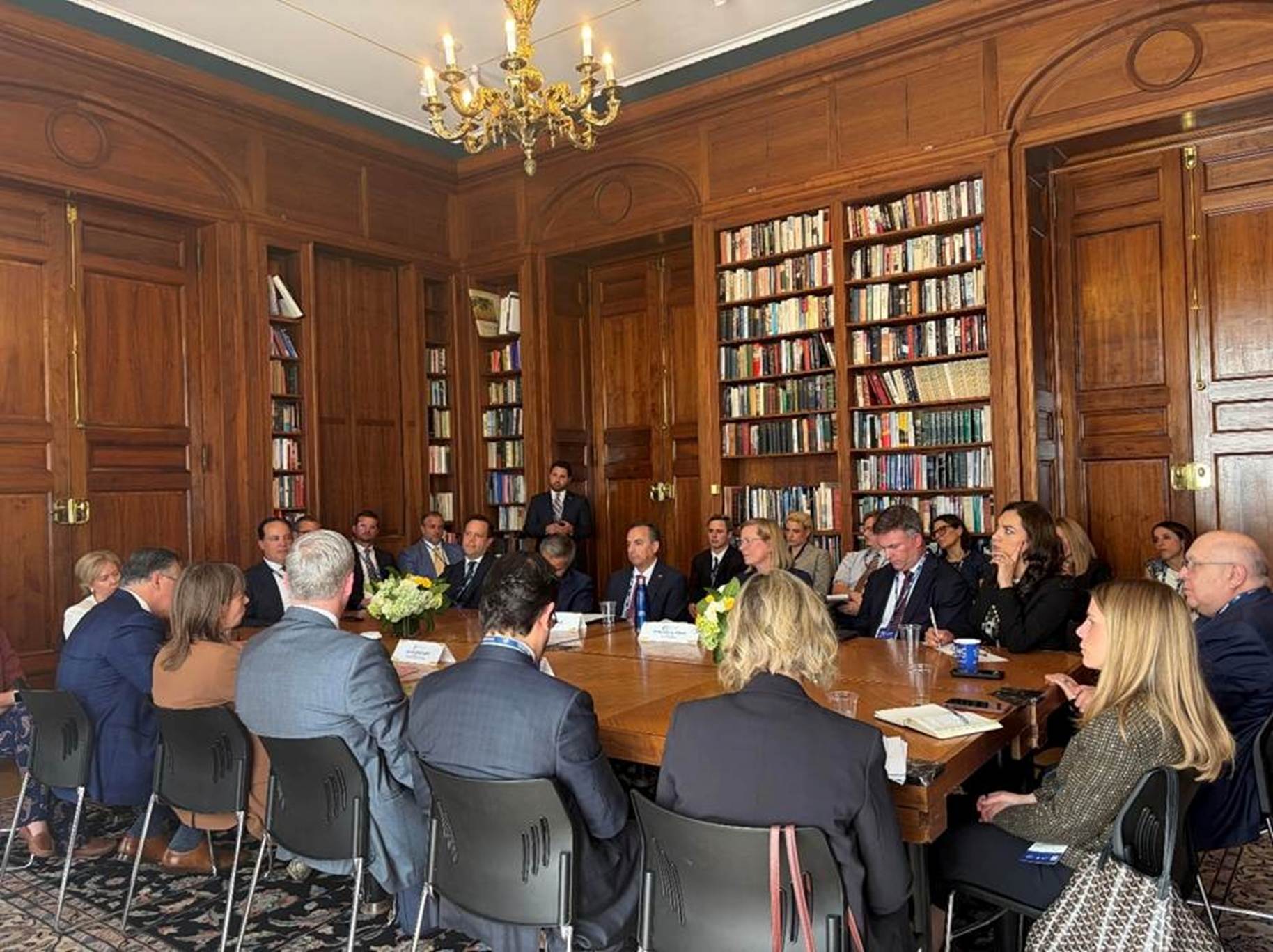 The image depicts a formal meeting room with a large, ornate wooden table surrounded by chairs, with people dressed in suits and ties, and a bookshelf filled with books in the background.
AI-generated content may be incorrect.